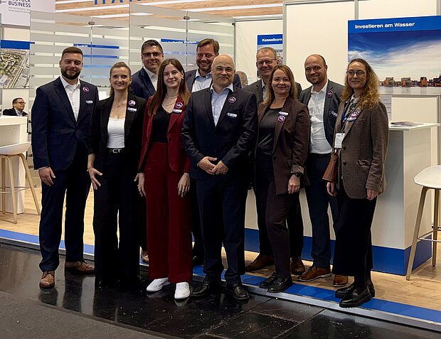 Zehn Mitarbeiter stehen vor einem Messestand und lächeln in die Kamera