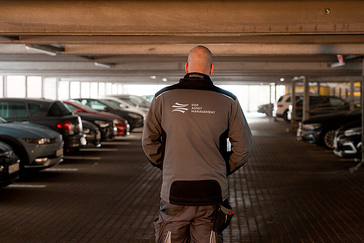 Infrastrukturelle Facility Services von DSR Asset Management Haustechniker steht mittig im Parkhaus, umgeben von Autos